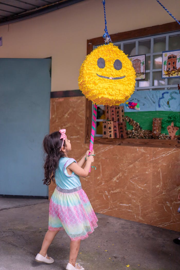 A young girl with long hair plays with a smiley face piñata indoors, in a colorful dress.